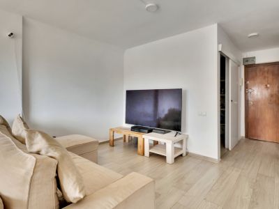 Helles Wohnzimmer mit beigem Ecksofa und TV auf hellen Holztischen. Der helle Laminatboden führt zur dunklen Holzeingangstür und einem offenen Regalbereich.