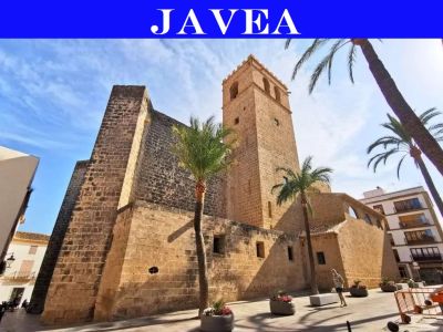 Historische Steinkirche mit Glockenturm in Jávea, mit robustem Mauerwerk und Strebepfeilern, auf einem sonnigen Platz mit Palmen.