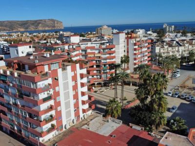 Luftaufnahme des Wohnkomplexes mit weißen Fassaden, rot-orangen Balkonen, Palmen und dem Mittelmeer im Hintergrund.