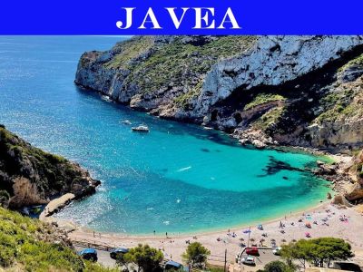 Luftaufnahme einer Bucht in Jávea mit klarem türkisfarbenem Wasser, einem Kiesstrand mit Badegästen und felsigen Klippen, die von mediterraner Vegetation bewachsen sind.