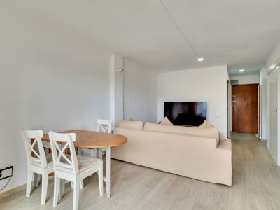 Offener Wohn- und Essbereich mit hellem Holztisch, weißen Stühlen und beigem Sofa. Ein Wandfernseher und heller Laminatboden führen zu einer dunklen Holzeingangstür.