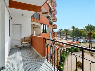 Sonniger Balkon mit blau-weißem geometrischem Fliesenboden, ausgestattet mit einem Korbstuhl und Abstellraumtür. Blick auf Palmen und den Wohnparkplatz.