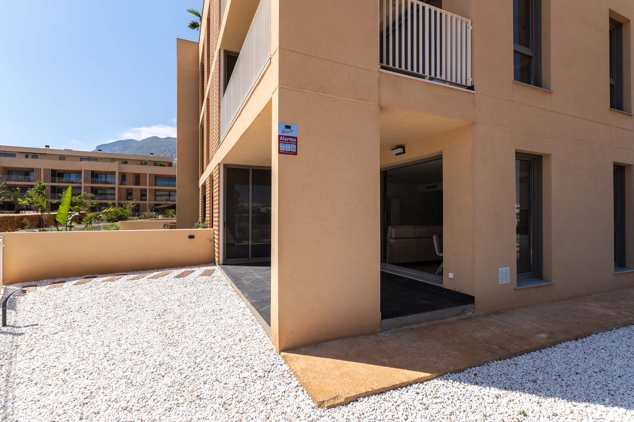 Außenansicht der Erdgeschosswohnung mit moderner ockerfarbener Fassade. Privater Vorgarten mit weißem Kies und überdachte Terrasse mit direktem Zugang, Bergblick im Hintergrund.