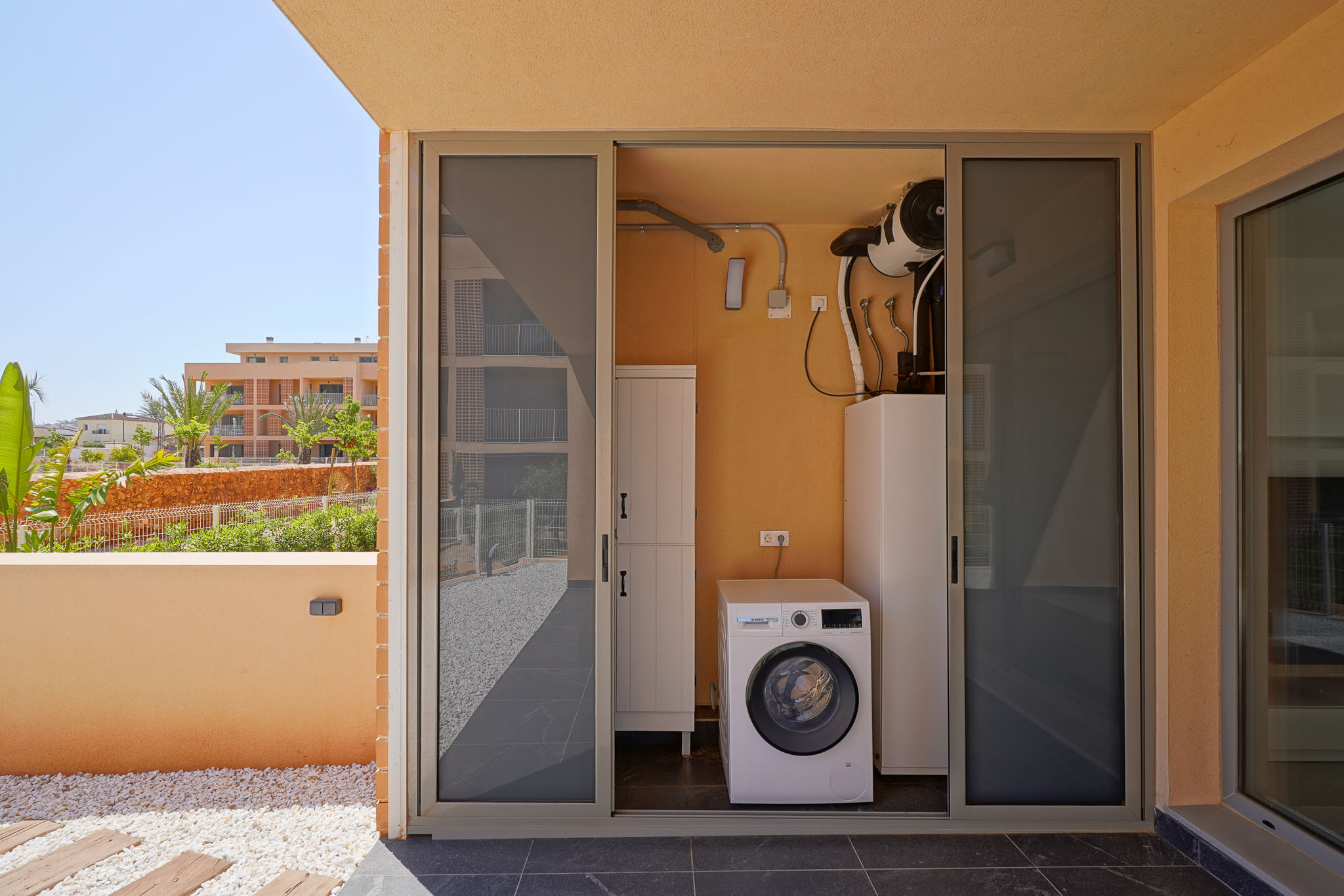 Außenwirtschaftsraum mit Bosch-Waschmaschine und Luftwärmepumpe, zugänglich von einer gefliesten Terrasse mit Zierkies und Gartenblick.