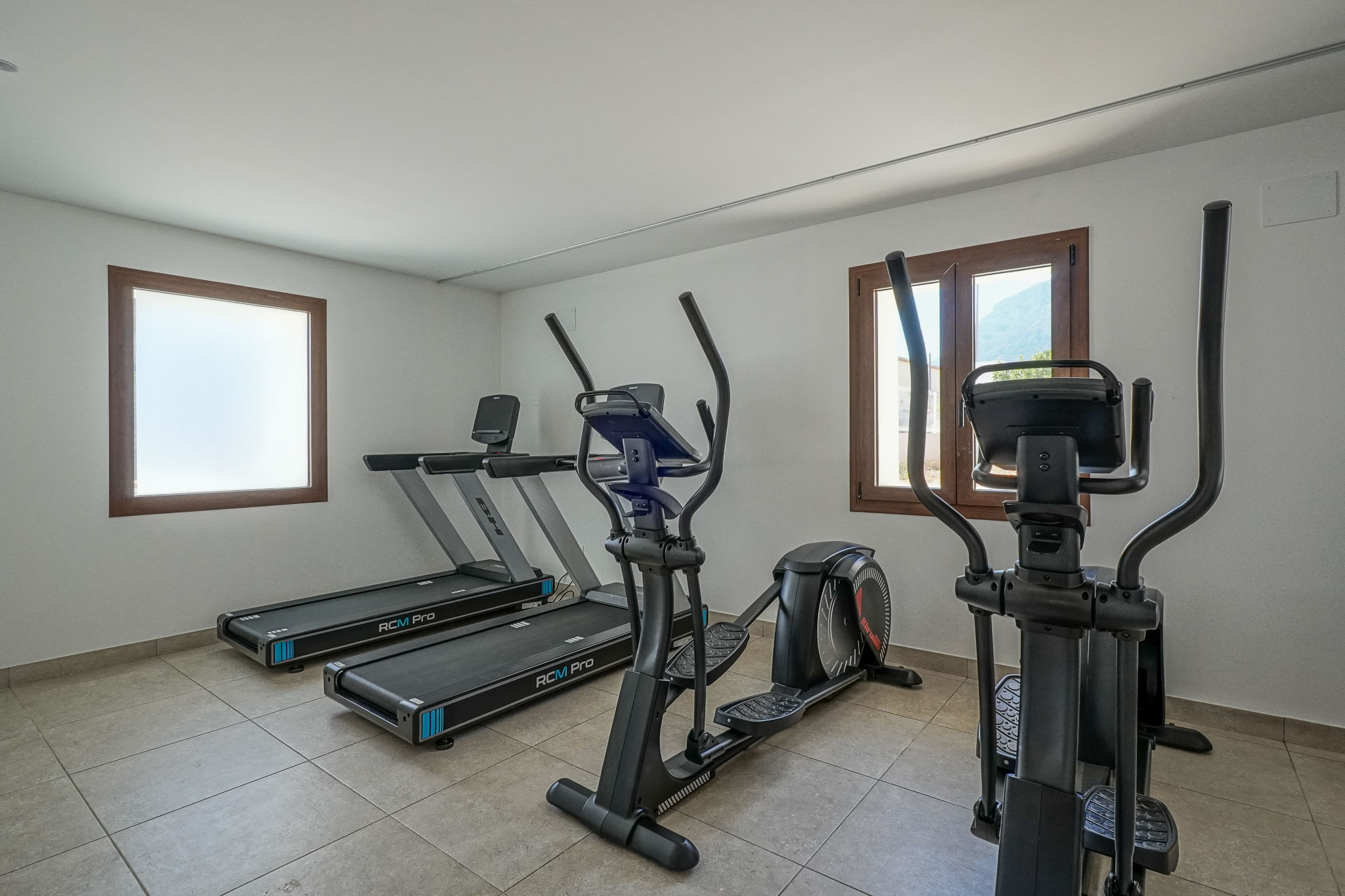 Fitnessraum der Wohnanlage mit zwei BH RCM Pro Laufbändern und zwei Ellipsentrainern. Er verfügt über einen Fliesenboden, weiße Wände und Holzfensterrahmen, eines davon mit Bergblick.