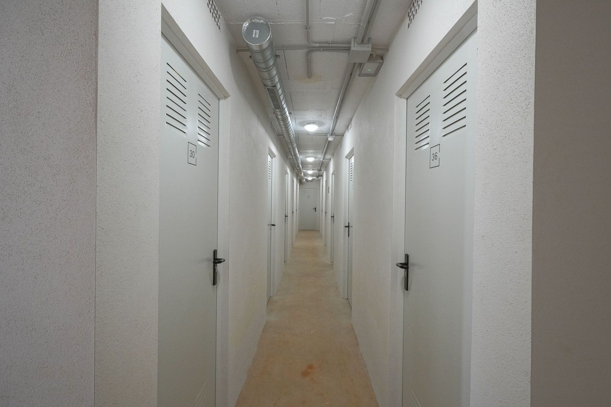 Funktionaler Korridor zu Lagereinheiten, mit nummerierten weißen Türen und Lüftungsschlitzen. Sichtbetondecke mit industriellen Lüftungskanälen und Einbauleuchten, ergänzt durch strukturierte Wände.