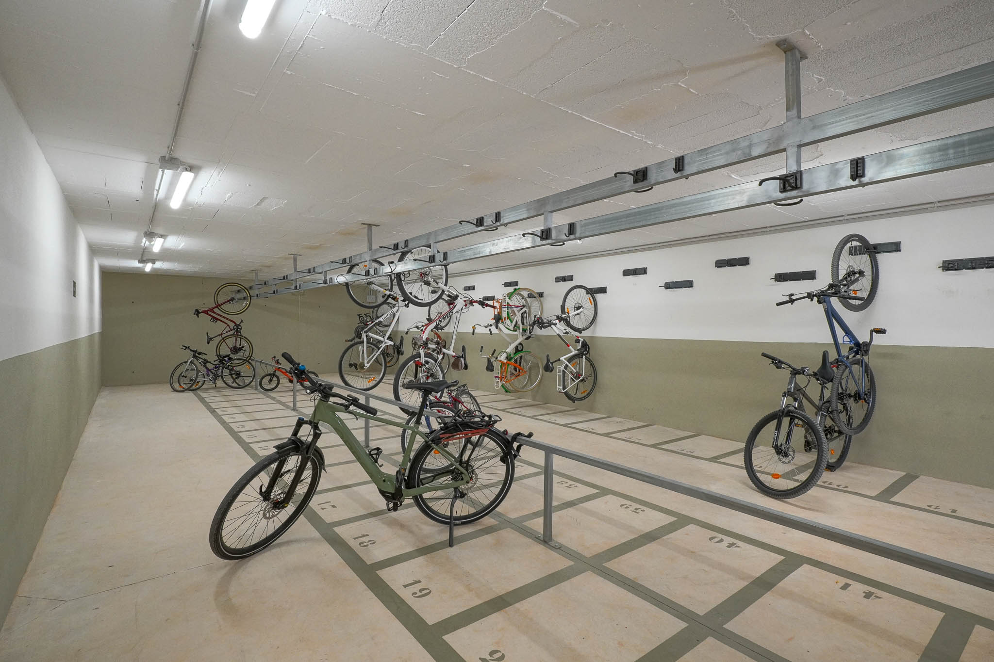 Geräumiger Gemeinschafts-Fahrradabstellbereich mit nummerierten Bodenplätzen und Wandhalterungen. Hell beleuchtet durch Deckenleuchten, bietet er sichere Aufbewahrung im Wohnkomplex.