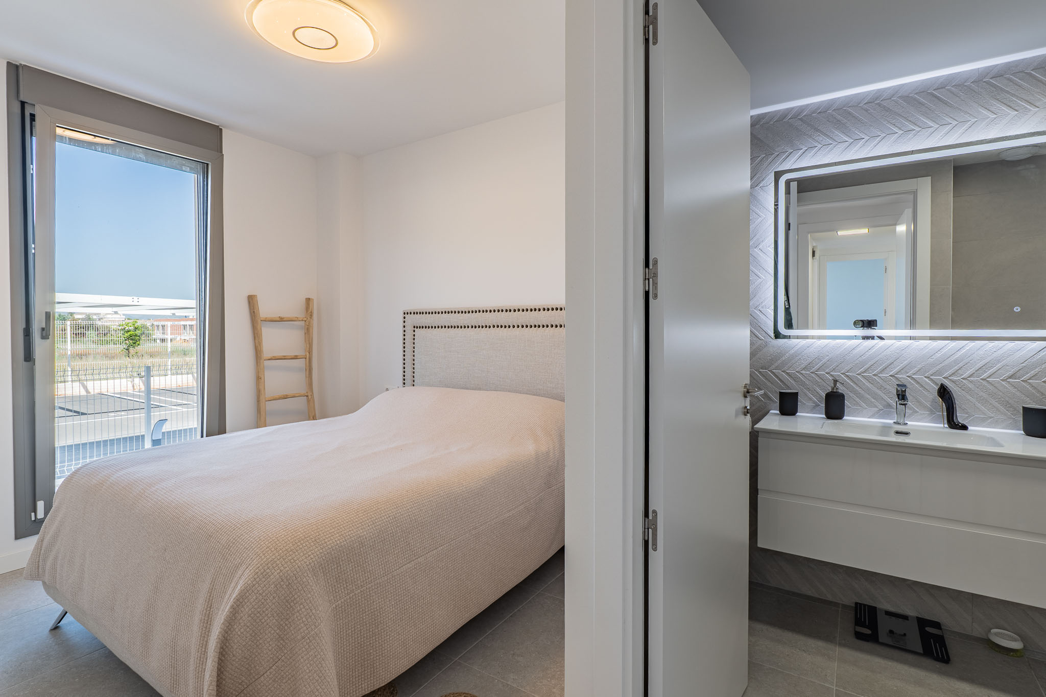 Geräumiges Schlafzimmer mit gepolstertem Kopfteilbett und direktem Zugang zur Außenterrasse. Ein en-suite Badezimmer mit schwebendem Waschtisch und beleuchtetem Spiegel ist sichtbar.