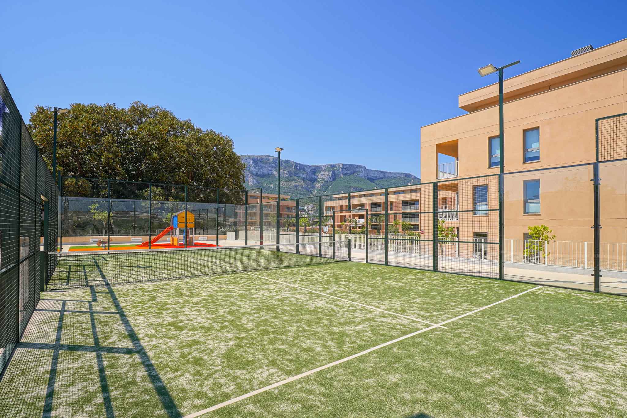 Grüner Kunstrasen-Padelplatz, eingezäunt und beleuchtet, neben einem Kinderspielplatz und modernen Wohngebäuden mit Bergblick.