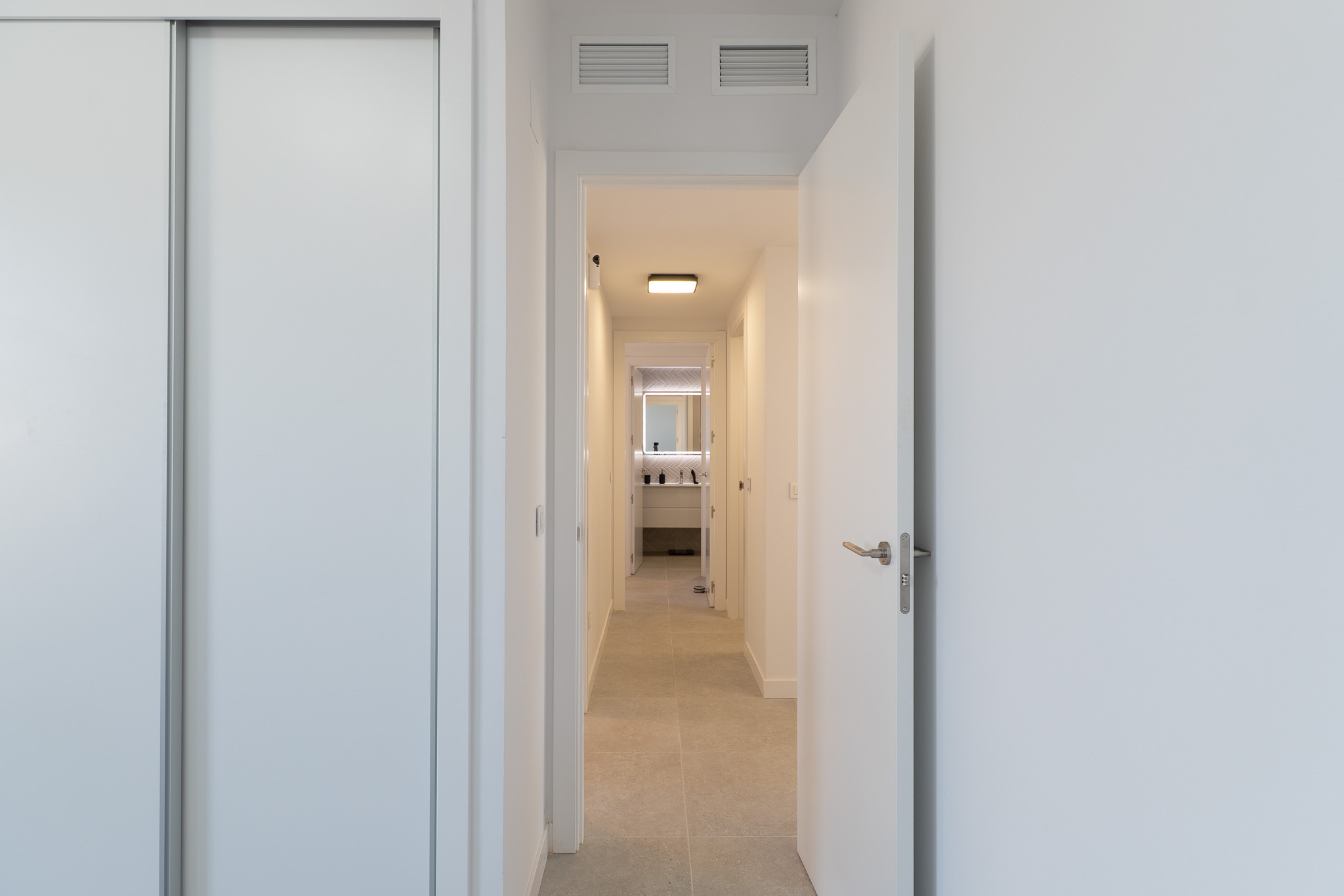 Heller Flur mit weißem Schiebetürenschrank und offener Tür. Helle Bodenfliesen führen zu einem modernen Badezimmer mit beleuchtetem Spiegel und stilvollem Waschtisch. Lüftungsgitter an der Decke.