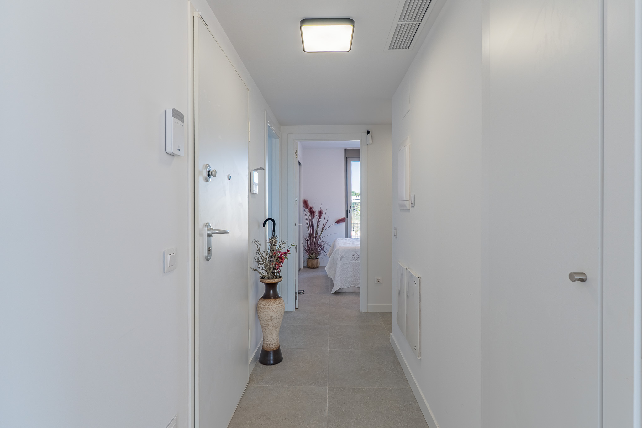 Heller, moderner Flur mit hellgrauen Fliesen und weißen Wänden. Ausgestattet mit Sicherheitspanel, moderner Deckenleuchte und Blick in ein Schlafzimmer.
