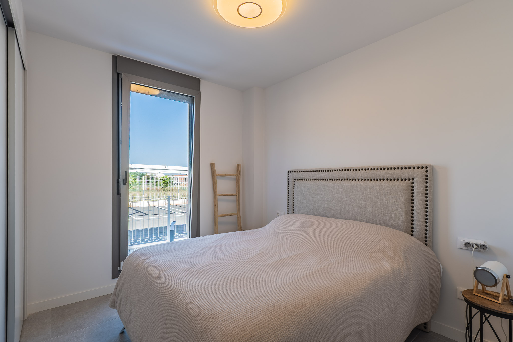 Helles Schlafzimmer mit Bett, genietetem Stoffkopfteil und Einbauschrank. Großes Fenster mit Zugang zum Außenbereich und grauen Fliesenboden.