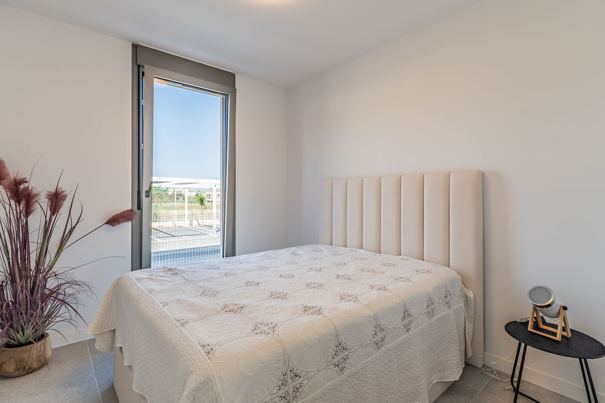 Helles Schlafzimmer mit Bett und hohem, gepolstertem beigem Kopfteil. Großes bodentiefes Fenster mit Außenblick. Graue Fliesen und eine Zierpflanze ergänzen den Raum.