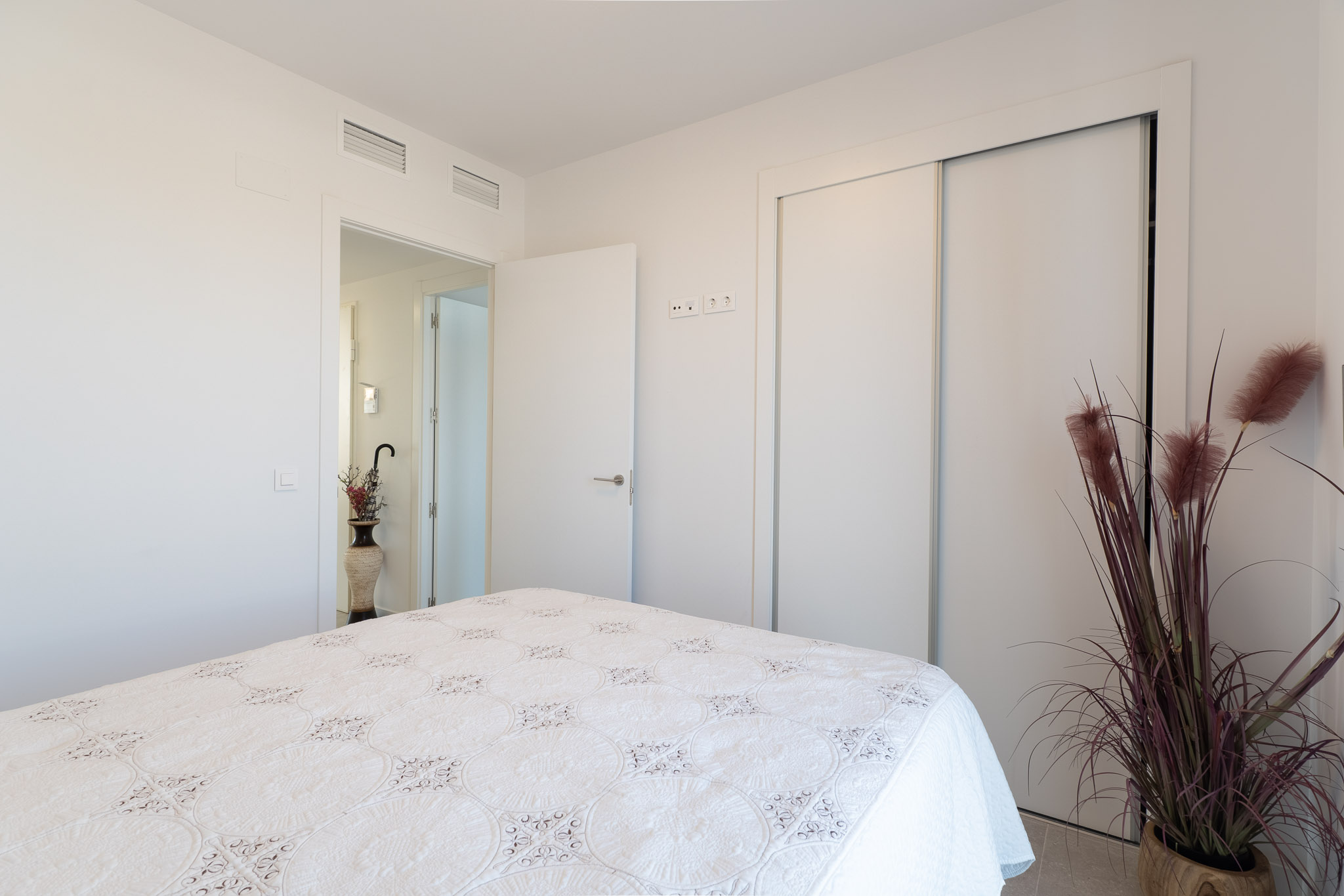 Helles Schlafzimmer mit Bett und weißer, gemusterter Tagesdecke. Es verfügt über einen integrierten Schiebeschrank und sichtbare Klimaanlagenlüftungsschlitze an der Decke. Eine Zierpflanze setzt Akzente.
