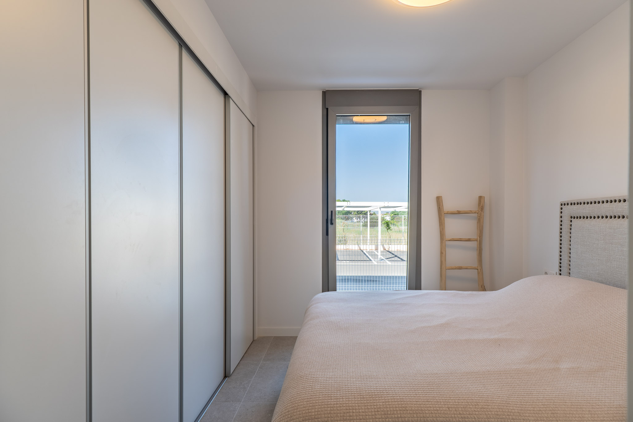 Helles Schlafzimmer mit weißem Einbauschrank und Schiebetüren, Fliesenboden und direktem Zugang zum Außenbereich durch eine Glastür. Bett mit gepolstertem Kopfteil.