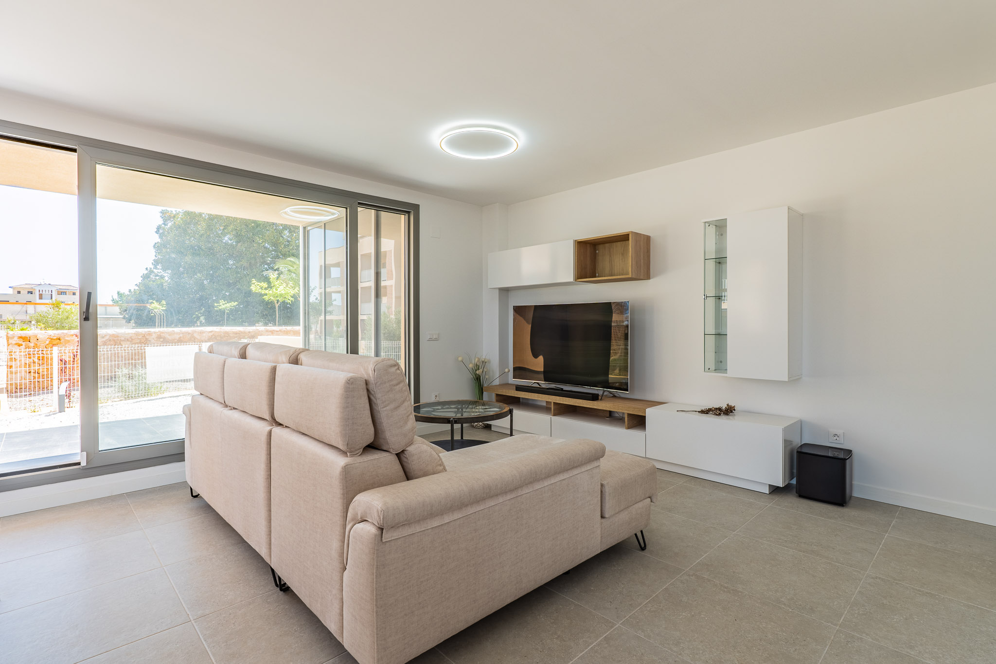 Helles Wohnzimmer mit beigem Ecksofa, hellgrauem Fliesenboden und großen Glasschiebetüren zum Außenbereich. Moderne TV-Möbelkombination mit weißen und Holzregalen.