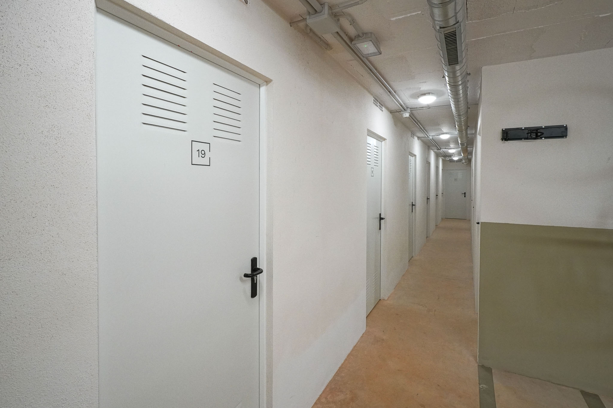 Langer Lagerflur mit weißen Türen, Lüftungsschlitzen und schwarzen Griffen. Die Decke zeigt sichtbare Lüftungskanäle und Einbauleuchten über einem Betonboden.