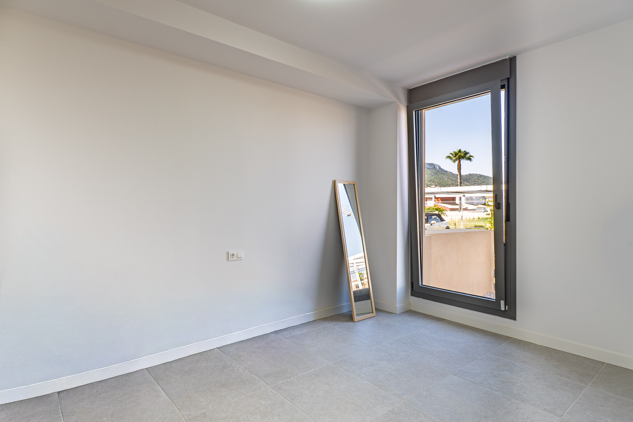Leerer Raum mit weißen Wänden und grauen Fliesen, bereit zur individuellen Gestaltung. Großes Fenster mit Blick auf eine Palme und Berge, das viel Tageslicht spendet.