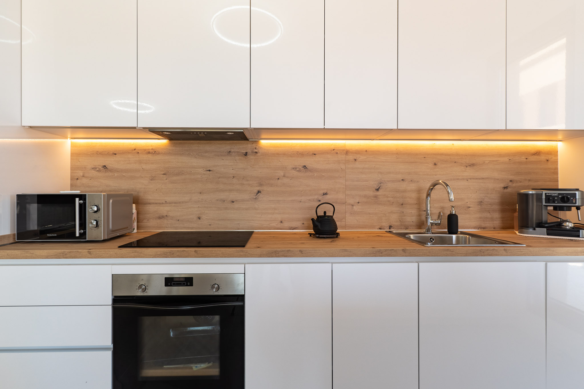 Moderne Küche mit glänzenden weißen Schränken, Arbeitsplatte und Rückwand in Holzoptik sowie warmer LED-Unterbauleuchte. Ausgestattet mit Backofen, Induktionskochfeld und Mikrowelle.