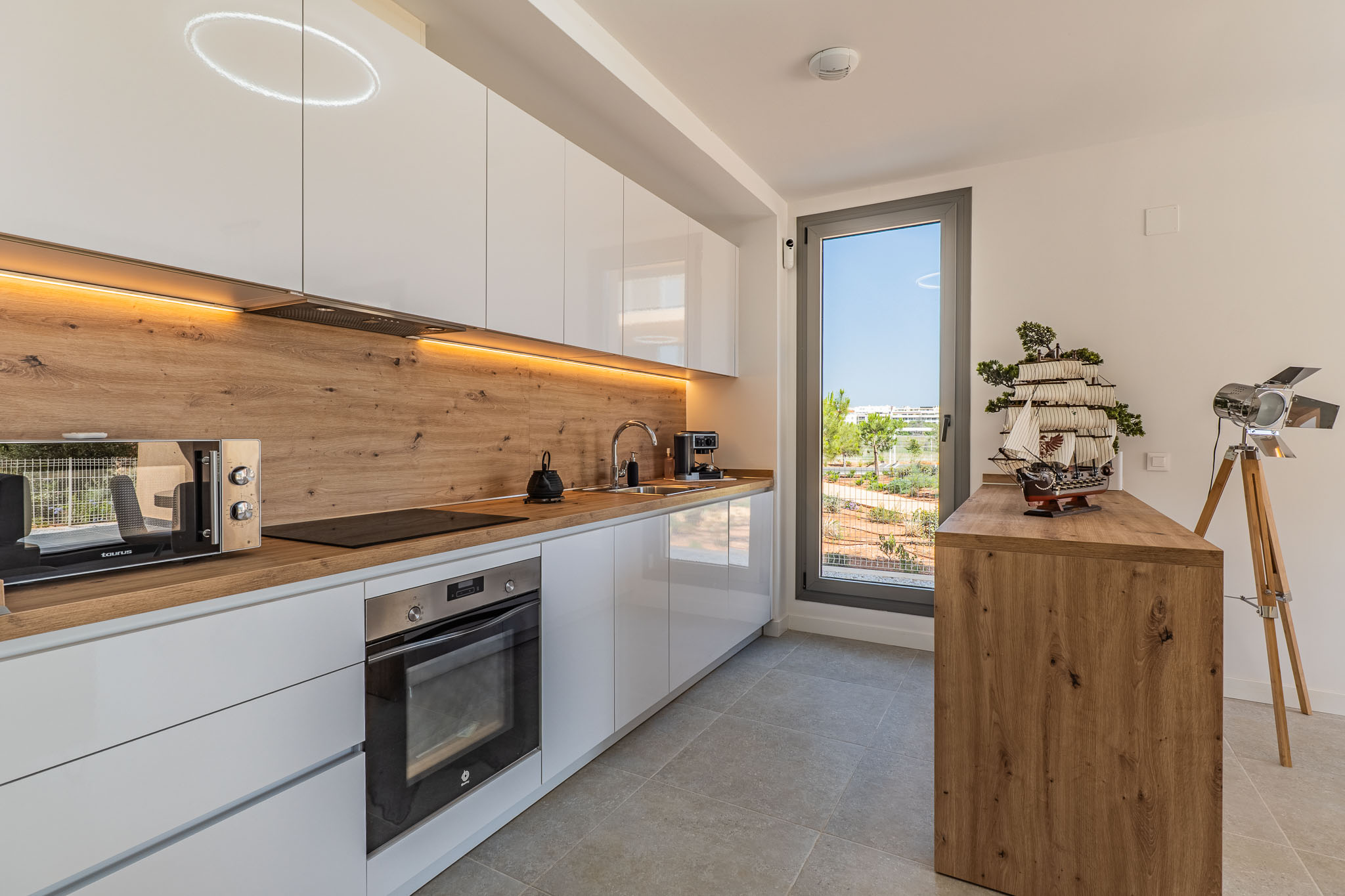 Moderne Küche mit hochglänzenden weißen Schränken, Arbeitsplatte und Spritzschutz in Holzoptik. Ausgestattet mit integrierten Geräten, LED-Beleuchtung und Fenster mit Blick ins Grüne.
