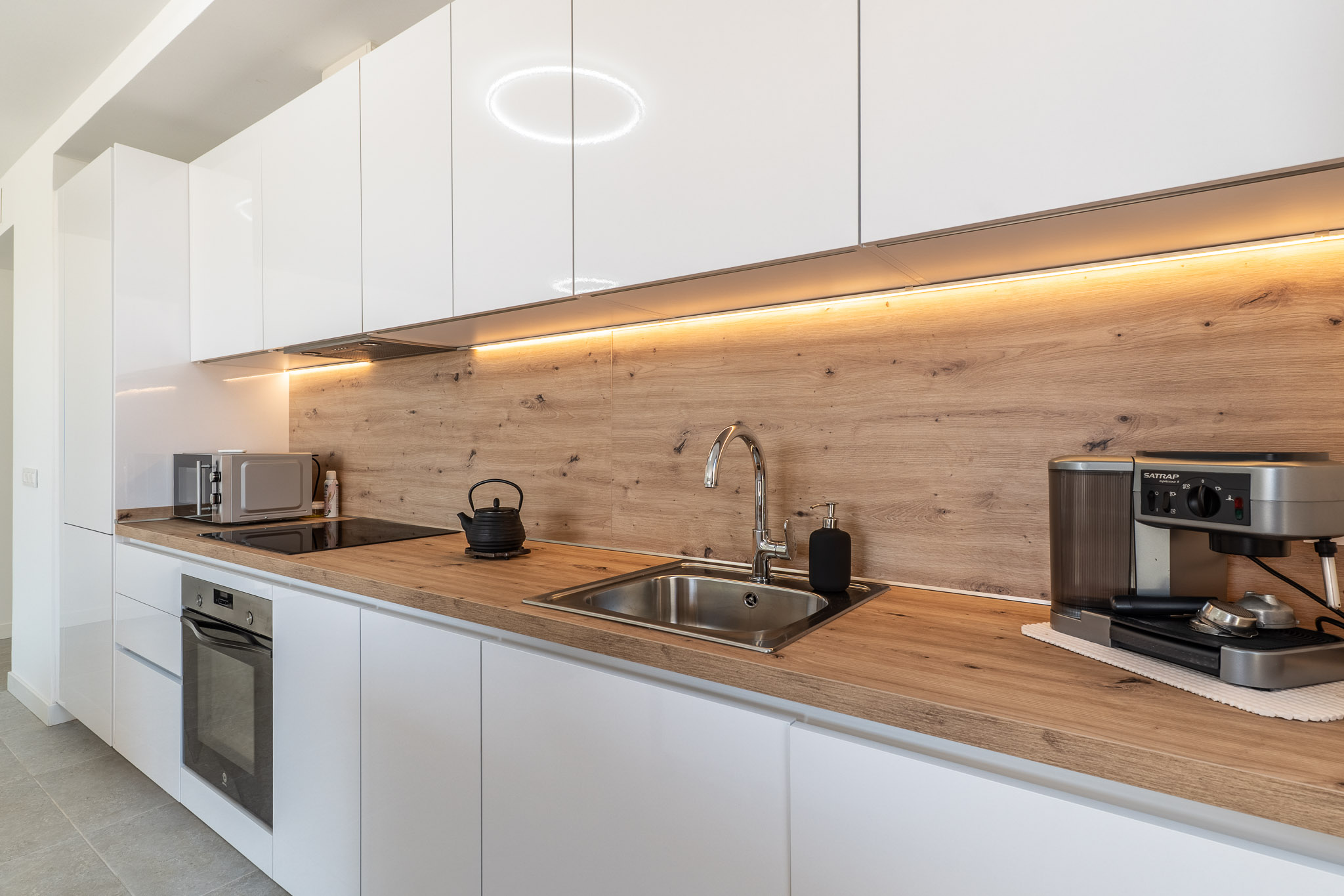 Moderne Küche mit hochglänzenden weißen Schränken, heller Holzoptik-Arbeitsplatte und passender Rückwand. Ausgestattet mit Einbaubackofen, Induktionskochfeld und Edelstahlspüle, beleuchtet durch LED-Unterbauleuchten.