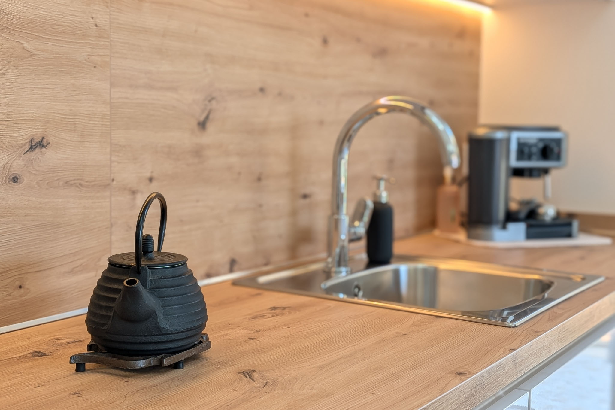 Moderne Küchenarbeitsplatte und Rückwand in heller Holzoptik. Edelstahlspüle mit Chromhahn, beleuchtet durch LED-Unterschrankleuchten. Eine Teekanne aus Gusseisen steht im Vordergrund.