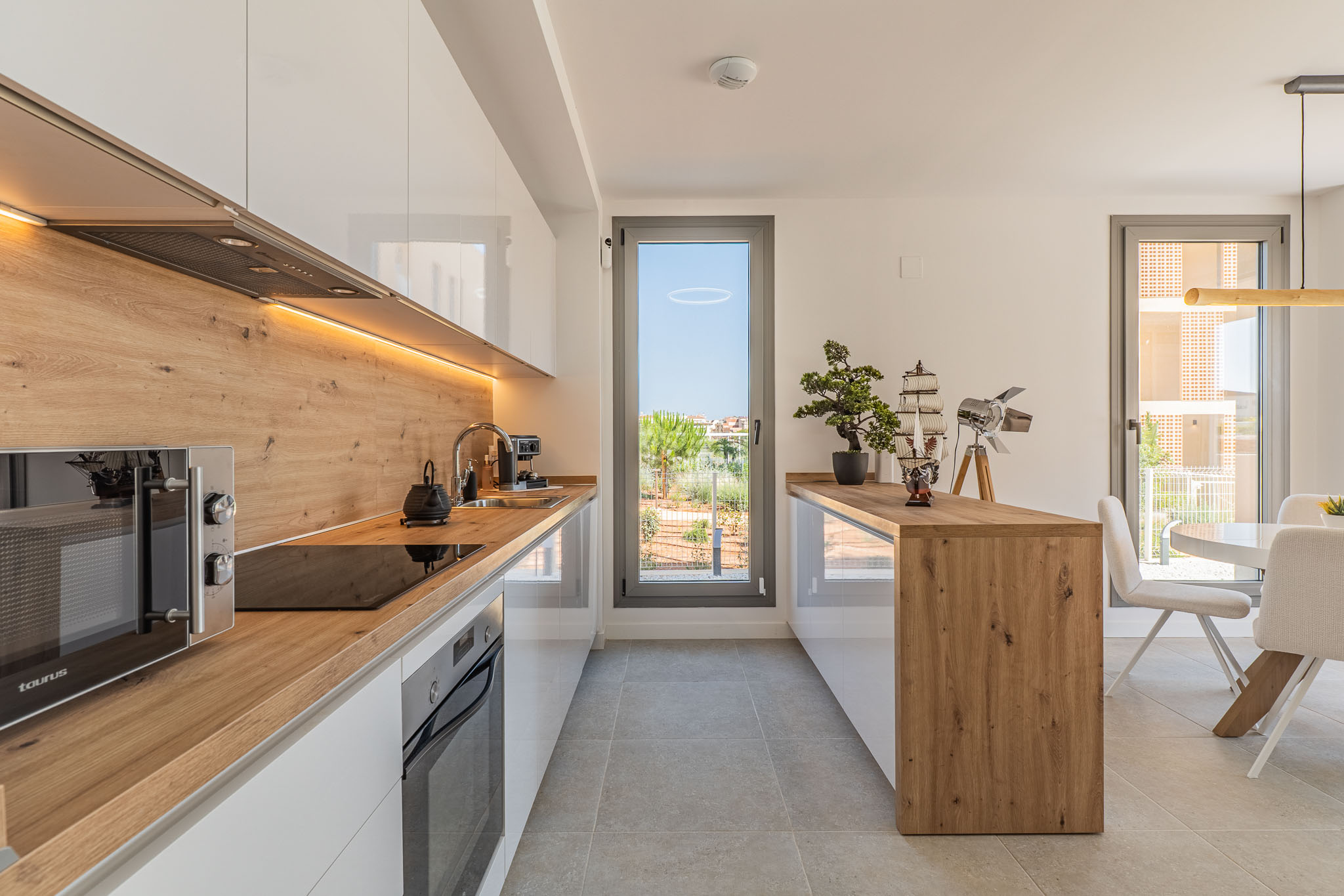 Moderne offene Küche mit glänzenden weißen Schränken, Arbeitsplatten und Spritzschutz in Holzoptik. Integrierte Geräte, LED-Beleuchtung und direkter Zugang zum Außenbereich.