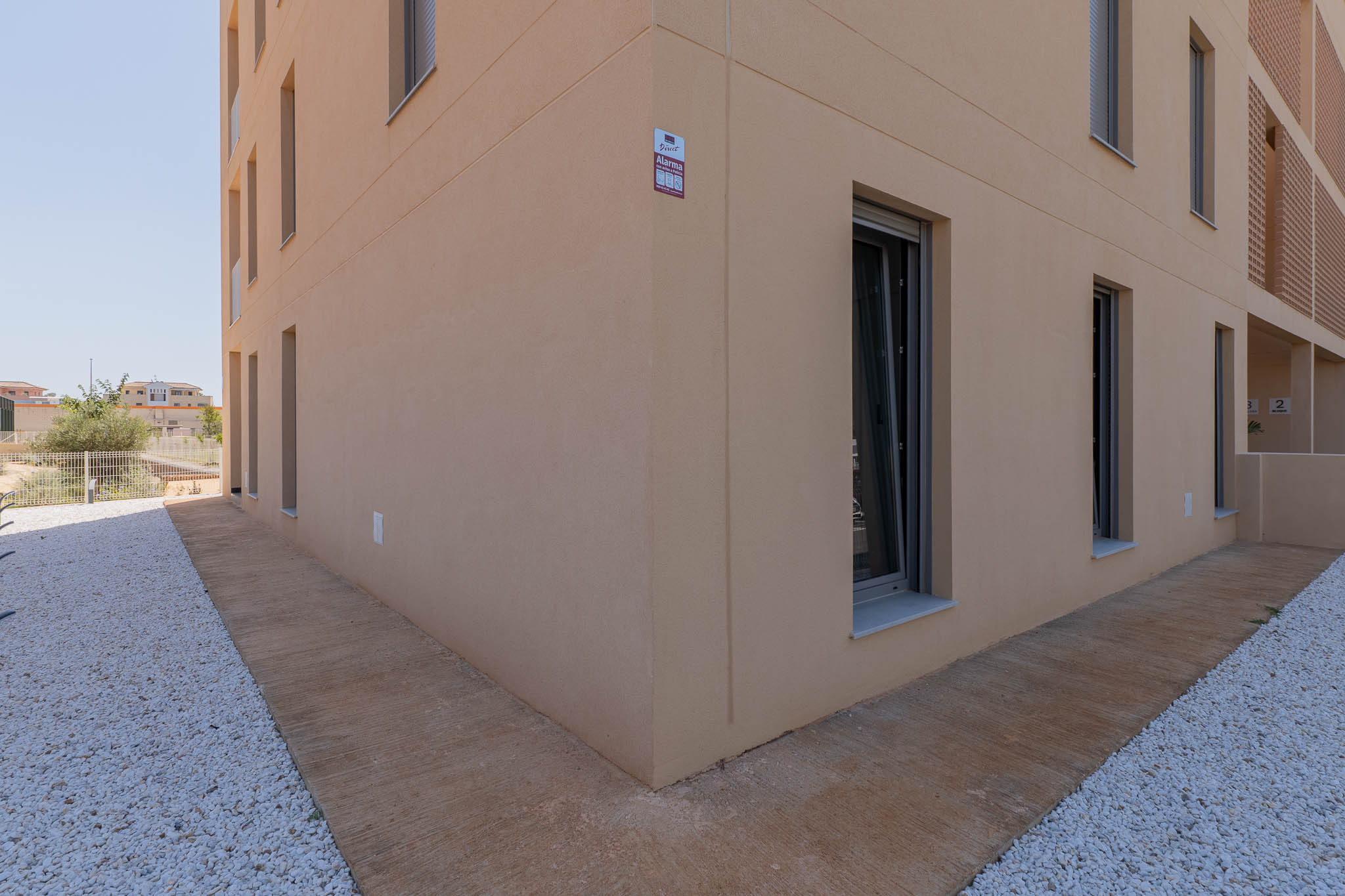 Moderner Gebäudeaußenbereich mit heller beigefarbener Putzfassade und dunkel gerahmten Fenstern. Der Boden zeigt einen strukturierten Betonweg neben einer weißen Kiesfläche, mit sichtbarer Alarmanlage.