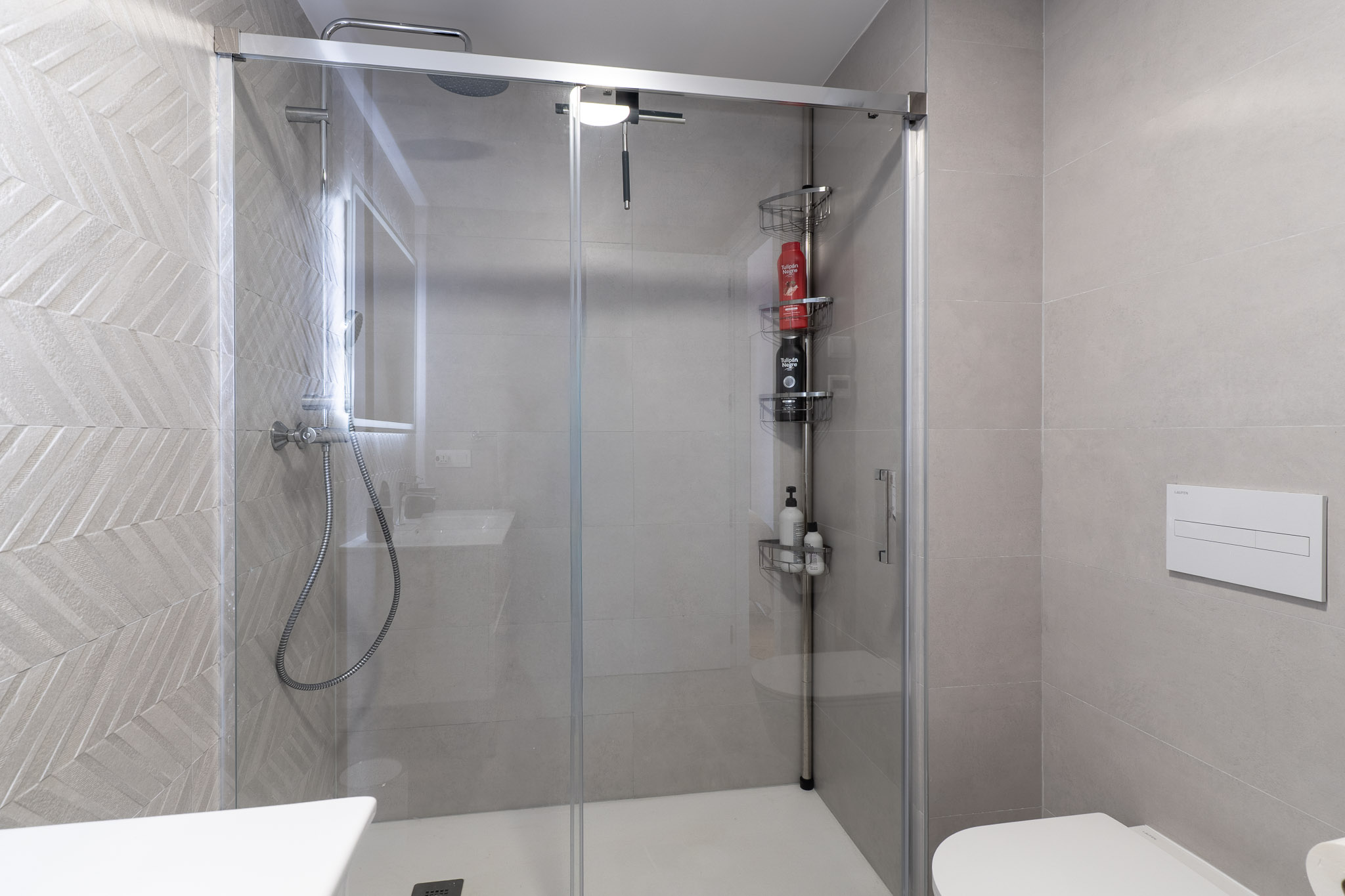 Modernes Badezimmer mit Glasduschkabine und weißer Duschwanne. Die Duschwand ist mit hellgrauen Fliesen im Fischgrätmuster versehen. Regendusche und wandhängendes WC sind sichtbar.