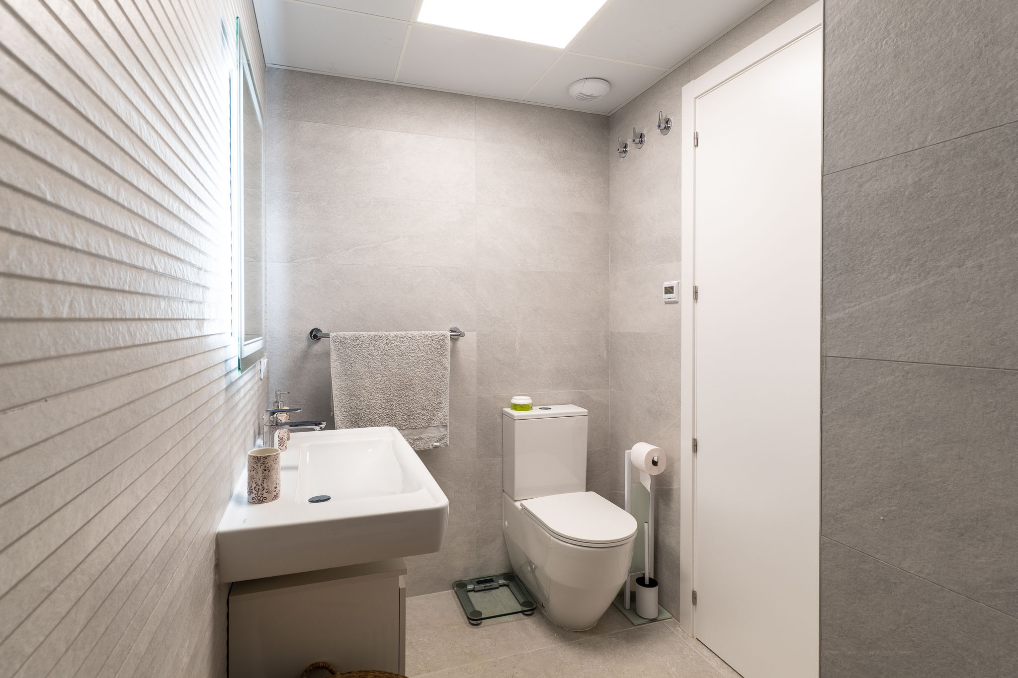 Modernes Badezimmer mit hellgrauen Großformatfliesen, Hänge-Waschbecken und WC. Ein beleuchteter Spiegel akzentuiert die strukturierte Wand.