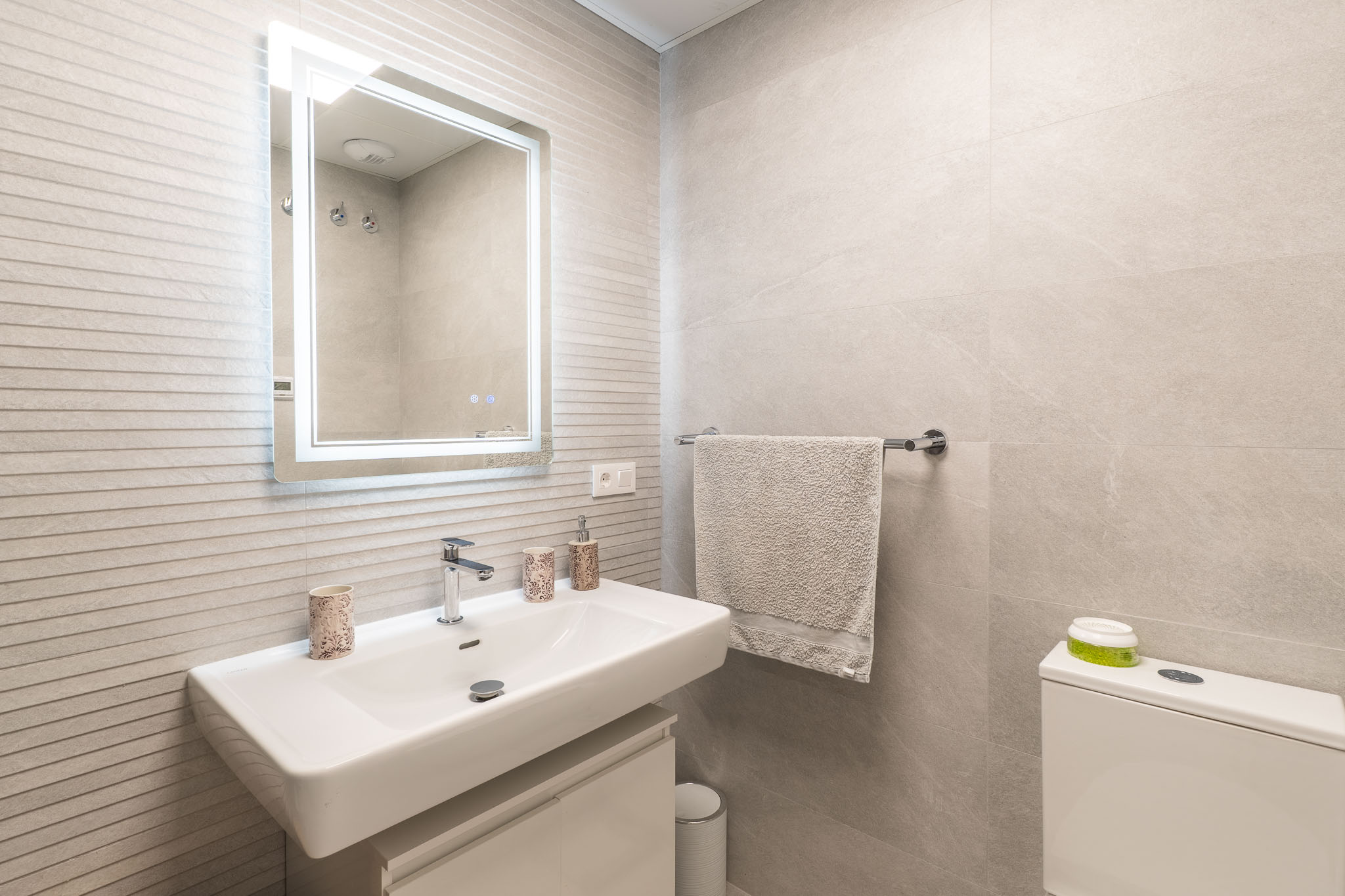 Modernes Badezimmer mit rechteckigem weißen Waschbecken, Wandarmatur, beleuchtetem Spiegel und hellgrauen strukturierten Wandfliesen.