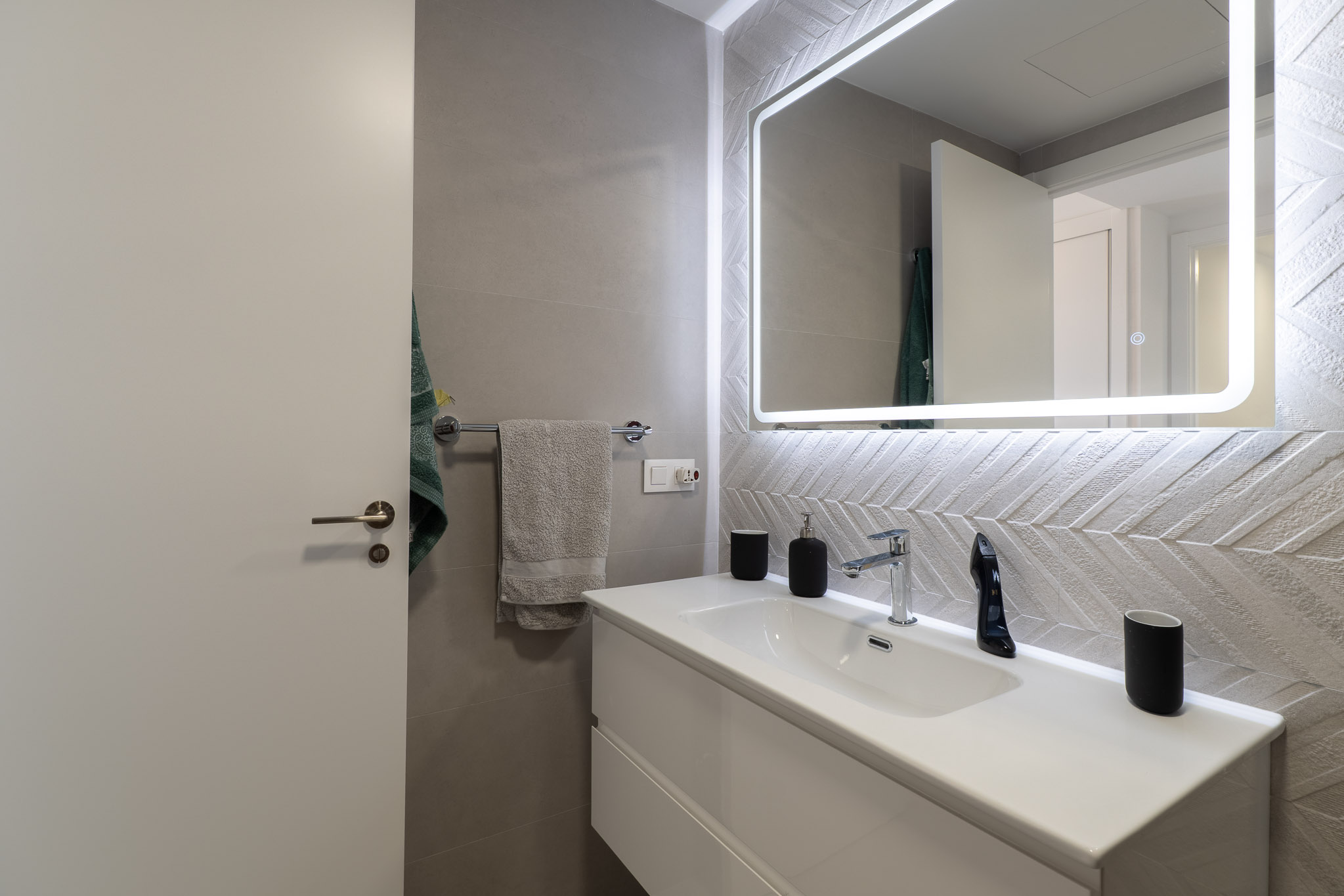 Modernes Badezimmer mit schwebendem Waschtisch, Chromarmatur und beleuchtetem Spiegel. Auffällig sind die strukturierten Wandfliesen im Fischgrätmuster.
