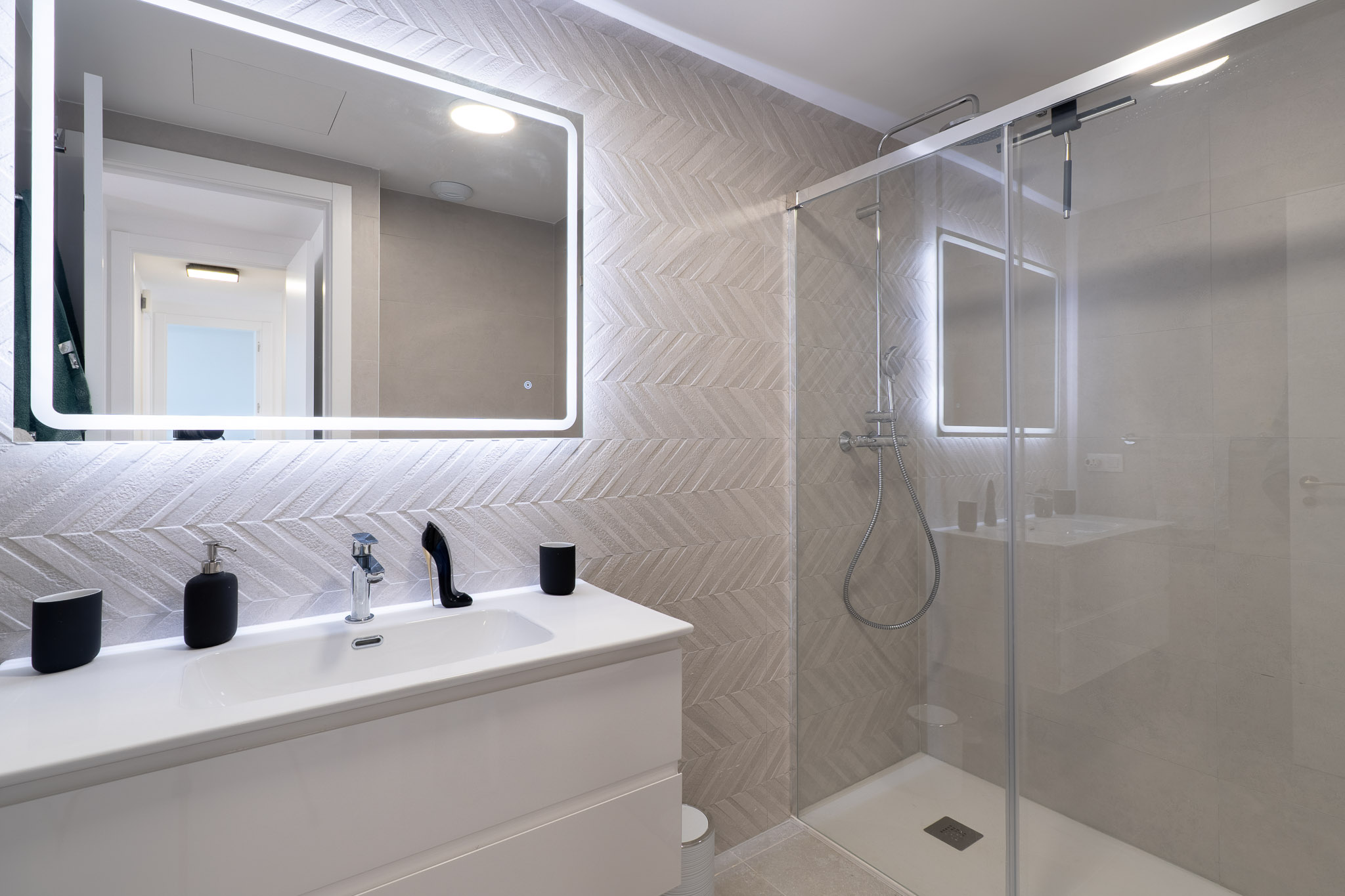 Modernes Badezimmer mit weißem Waschtisch, beleuchtetem Spiegel und Wandfliesen im Fischgrätmuster. Die Glasdusche verfügt über eine Regendusche und Chromarmaturen.