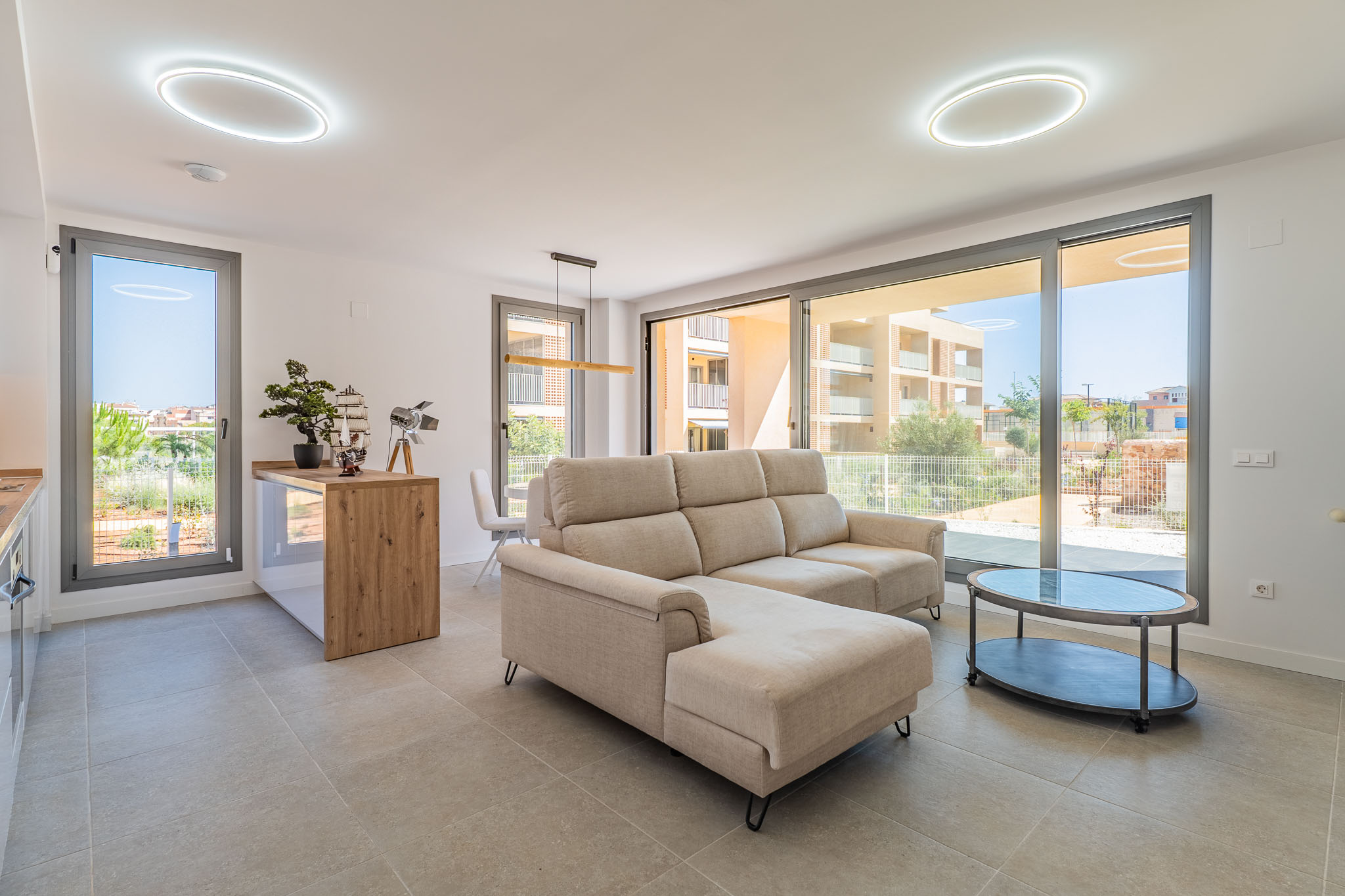 Offener Wohn- und Essbereich mit beigem Ecksofa und ovalem Couchtisch. Große Glasfronten sorgen für viel Tageslicht und Blick auf die Wohnanlage. Offene Küche mit Holztheke.