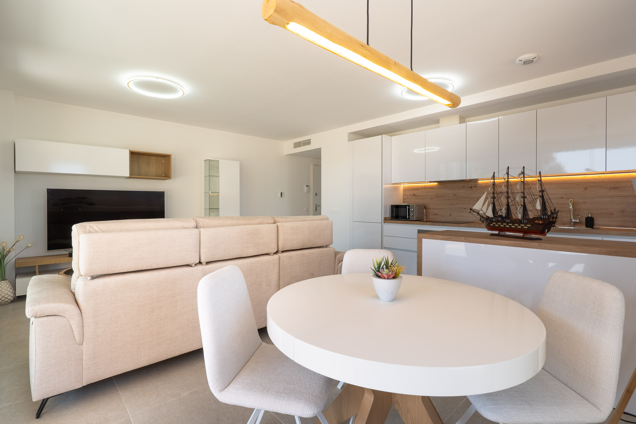 Offener Wohn- und Essbereich mit integrierter Küche. Helles Sofa, weißer ovaler Esstisch und moderne Küche mit hochglänzenden weißen Schränken, Holzdetails und indirekter Beleuchtung.