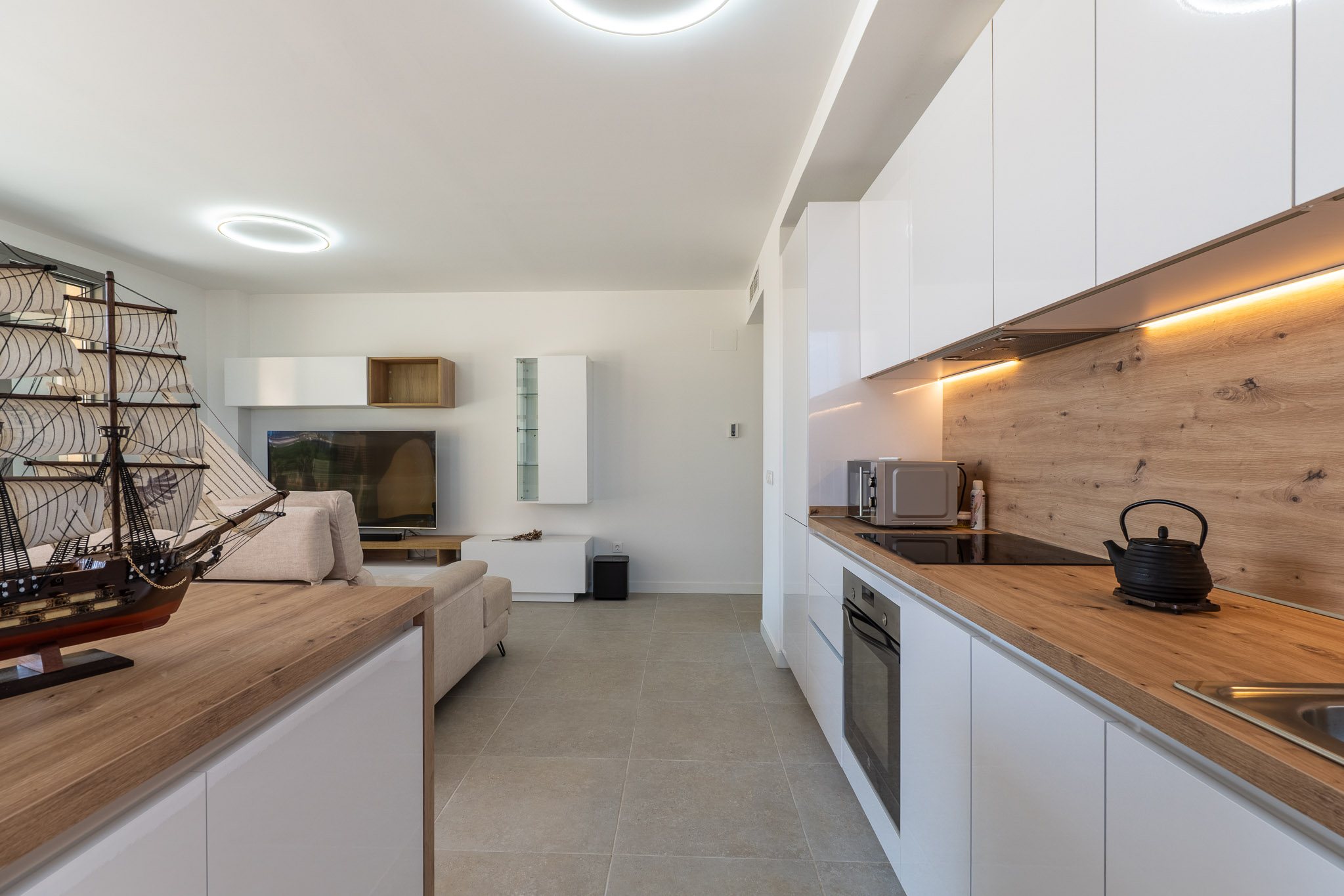 Offener Wohn- und Essbereich mit moderner Küche. Weiße Hochglanzschränke, helle Arbeitsplatten in Holzoptik und integrierte Geräte werden durch warmes LED-Licht und graue Fliesenböden betont.