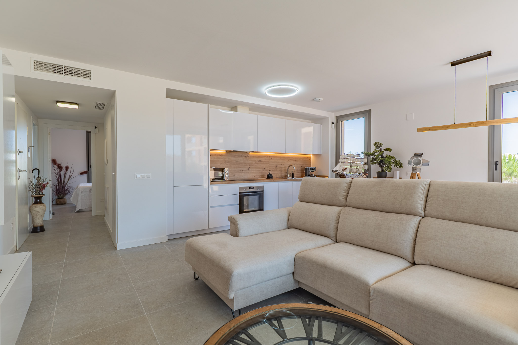 Offener Wohn- und Küchenbereich mit beigem Ecksofa, modernen weißen Hochglanzschränken und heller Holzrückwand. Fliesenboden und Tageslicht durch große Fenster.
