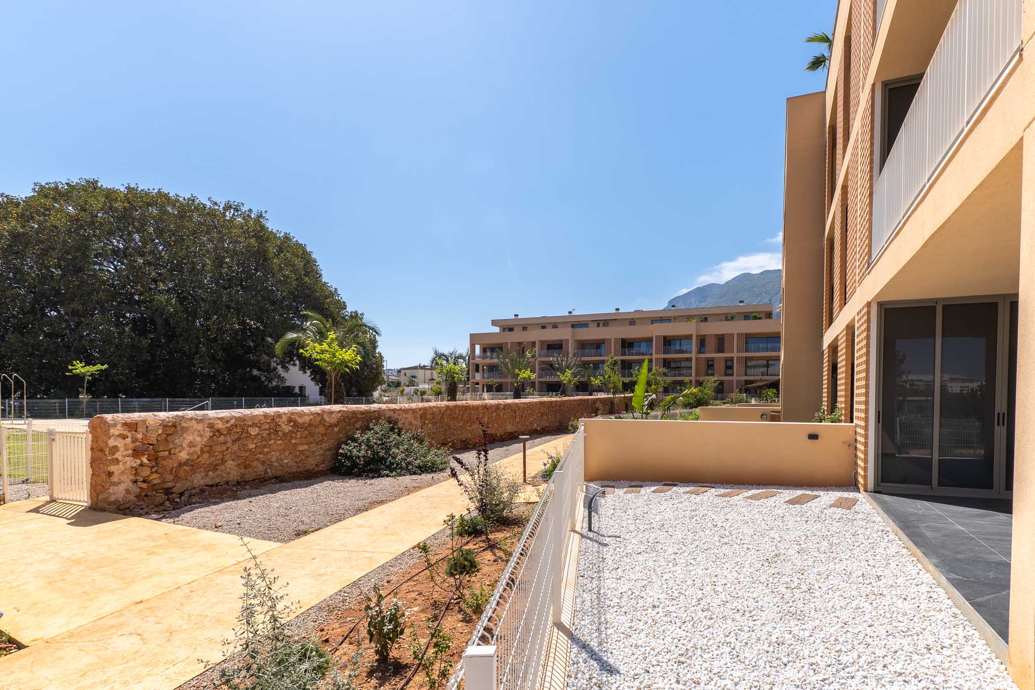 Privater Erdgeschossgarten mit weißem Kies und Trittsteinen, umzäunt von einem niedrigen weißen Zaun und einer Steinmauer. Blick auf moderne Gebäude und entfernte Berge.