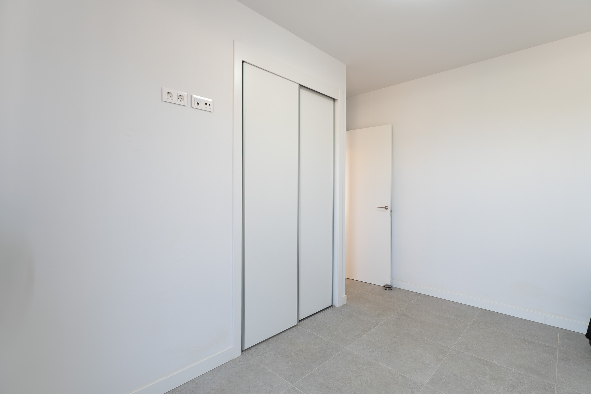 Schlafzimmer mit großem Einbauschrank mit weißen Schiebetüren und hellem Fliesenboden. Moderne Strom- und Datenanschlüsse sind vorhanden.