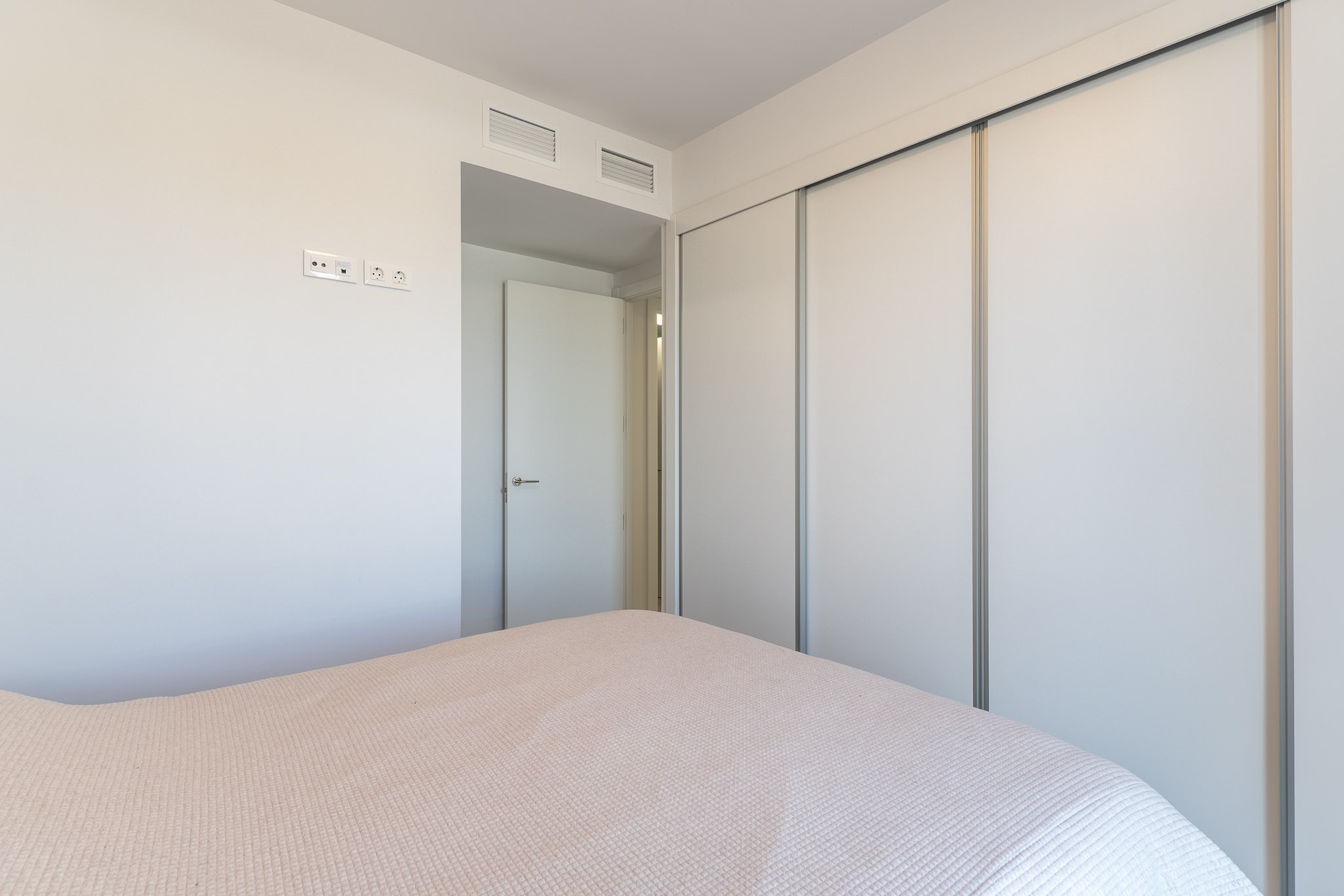 Schlafzimmeransicht mit einem geräumigen Einbauschrank mit weißen Schiebetüren. Die Wand zeigt moderne Steckdosen und integrierte Lüftungsgitter über der angrenzenden Tür.