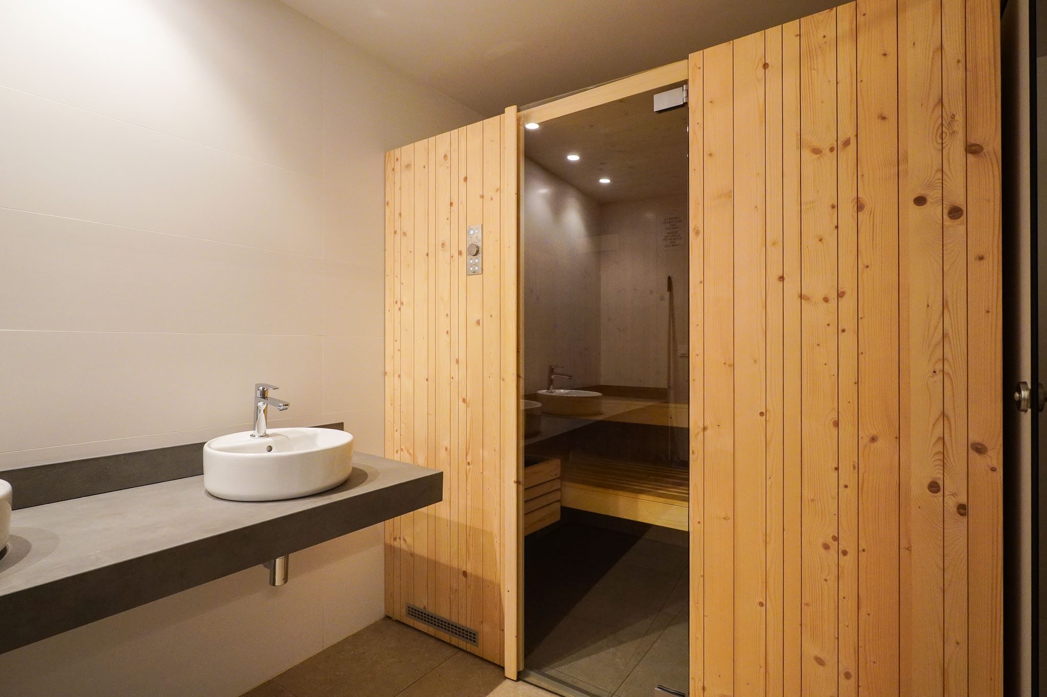 Umkleidebereich mit einem eleganten, schwebenden Waschtisch und Aufsatzwaschbecken sowie dem Holztür-Eingang zur Sauna mit Glaspaneel.
