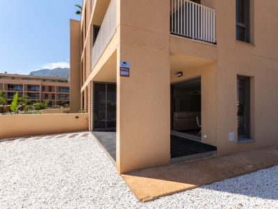 Außenansicht der Erdgeschosswohnung mit moderner ockerfarbener Fassade. Privater Vorgarten mit weißem Kies und überdachte Terrasse mit direktem Zugang, Bergblick im Hintergrund.