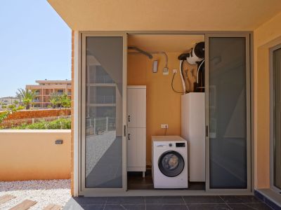 Außenwirtschaftsraum mit Bosch-Waschmaschine und Luftwärmepumpe, zugänglich von einer gefliesten Terrasse mit Zierkies und Gartenblick.