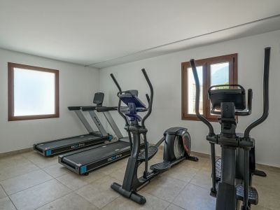 Fitnessraum der Wohnanlage mit zwei BH RCM Pro Laufbändern und zwei Ellipsentrainern. Er verfügt über einen Fliesenboden, weiße Wände und Holzfensterrahmen, eines davon mit Bergblick.