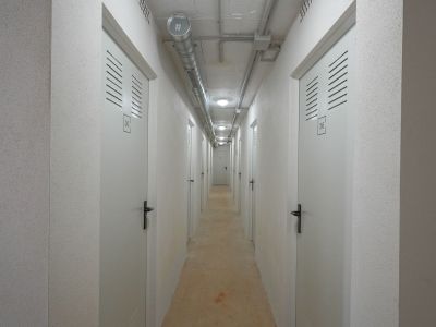 Funktionaler Korridor zu Lagereinheiten, mit nummerierten weißen Türen und Lüftungsschlitzen. Sichtbetondecke mit industriellen Lüftungskanälen und Einbauleuchten, ergänzt durch strukturierte Wände.