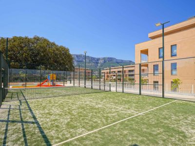Grüner Kunstrasen-Padelplatz, eingezäunt und beleuchtet, neben einem Kinderspielplatz und modernen Wohngebäuden mit Bergblick.