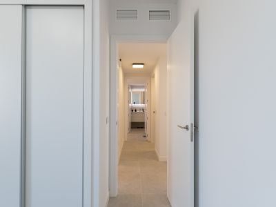Heller Flur mit weißem Schiebetürenschrank und offener Tür. Helle Bodenfliesen führen zu einem modernen Badezimmer mit beleuchtetem Spiegel und stilvollem Waschtisch. Lüftungsgitter an der Decke.