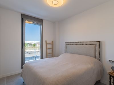 Helles Schlafzimmer mit Bett, genietetem Stoffkopfteil und Einbauschrank. Großes Fenster mit Zugang zum Außenbereich und grauen Fliesenboden.