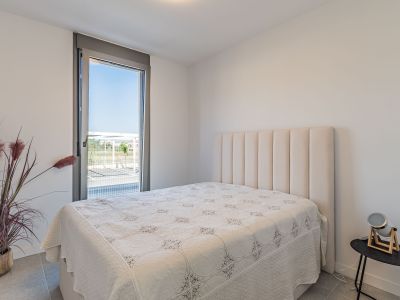 Helles Schlafzimmer mit Bett und hohem, gepolstertem beigem Kopfteil. Großes bodentiefes Fenster mit Außenblick. Graue Fliesen und eine Zierpflanze ergänzen den Raum.