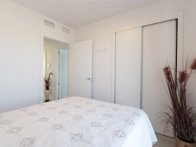 Helles Schlafzimmer mit Bett und weißer, gemusterter Tagesdecke. Es verfügt über einen integrierten Schiebeschrank und sichtbare Klimaanlagenlüftungsschlitze an der Decke. Eine Zierpflanze setzt Akzente.